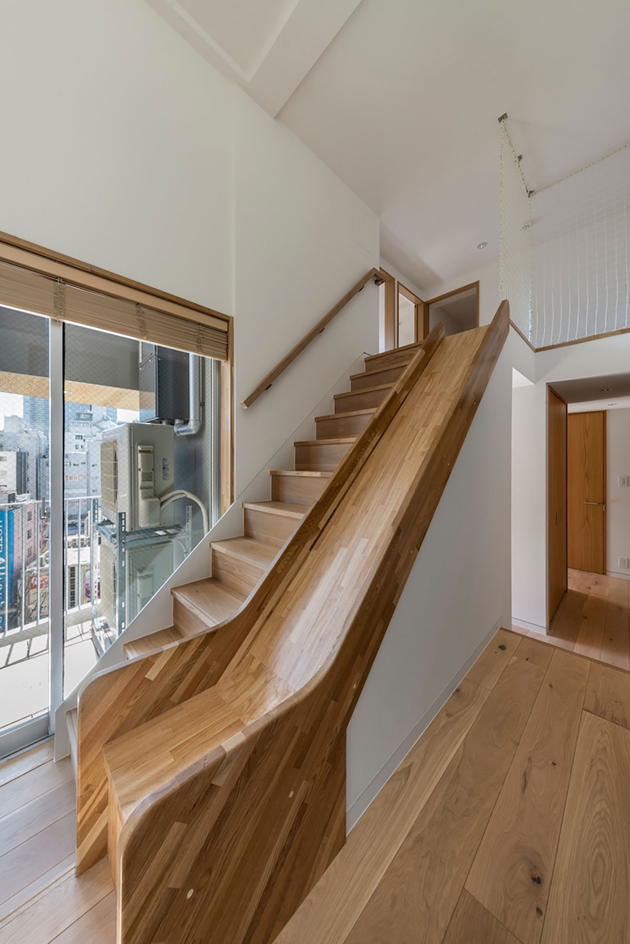 A modern interior with wooden stairs and an adjacent built-in wooden slide, next to large windows overlooking a cityscape.
