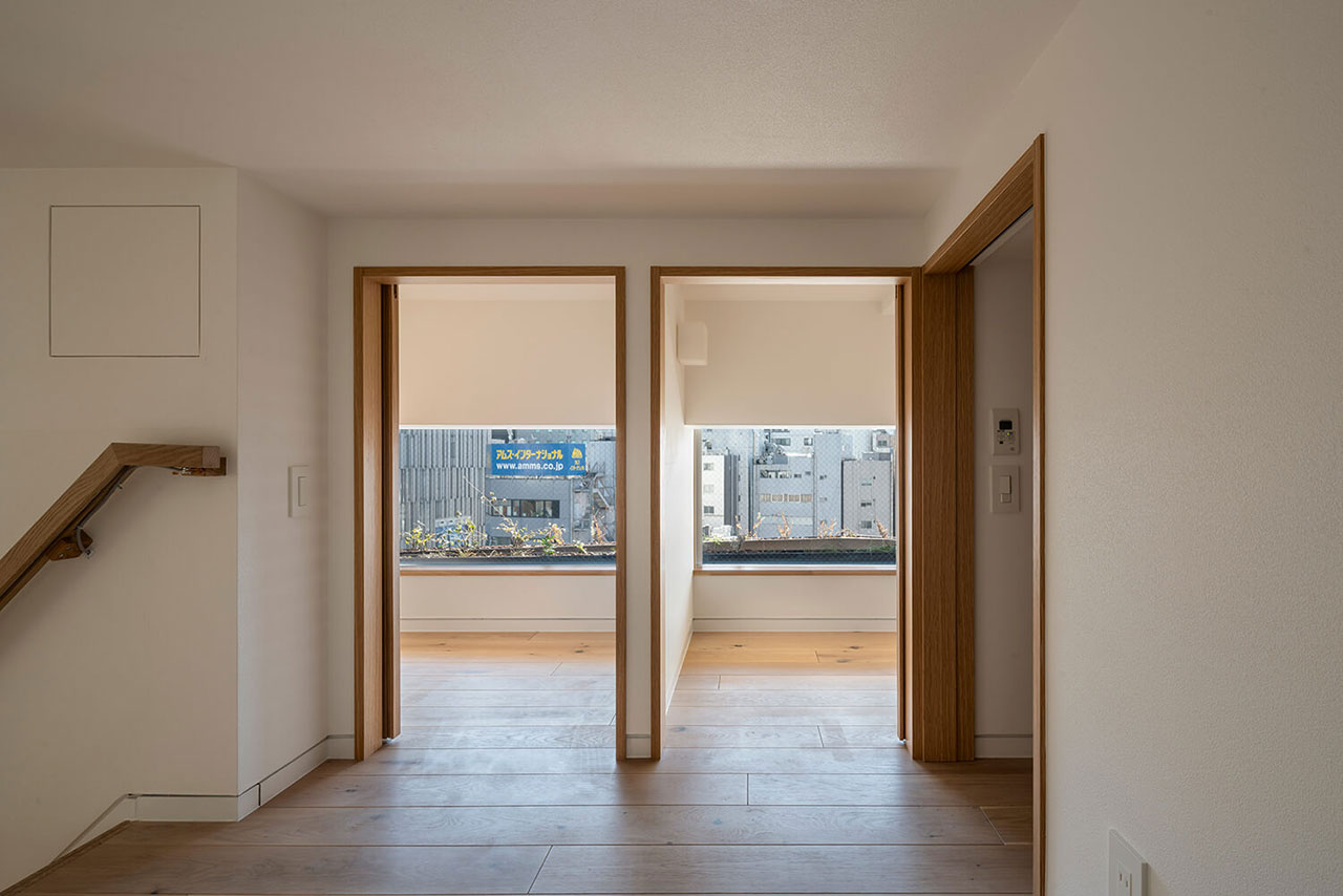 A modern, minimalist interior with light wood flooring, two doorways leading to rooms with large windows, and a partial staircase on the left.