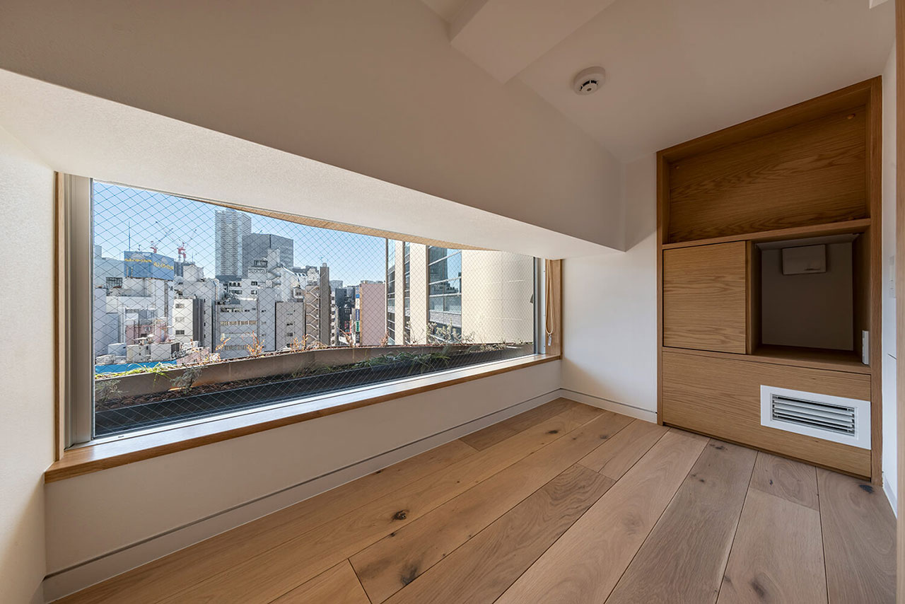 Small, empty room with a wide window overlooking city buildings, light wood flooring, and built-in wooden storage units on the right wall.