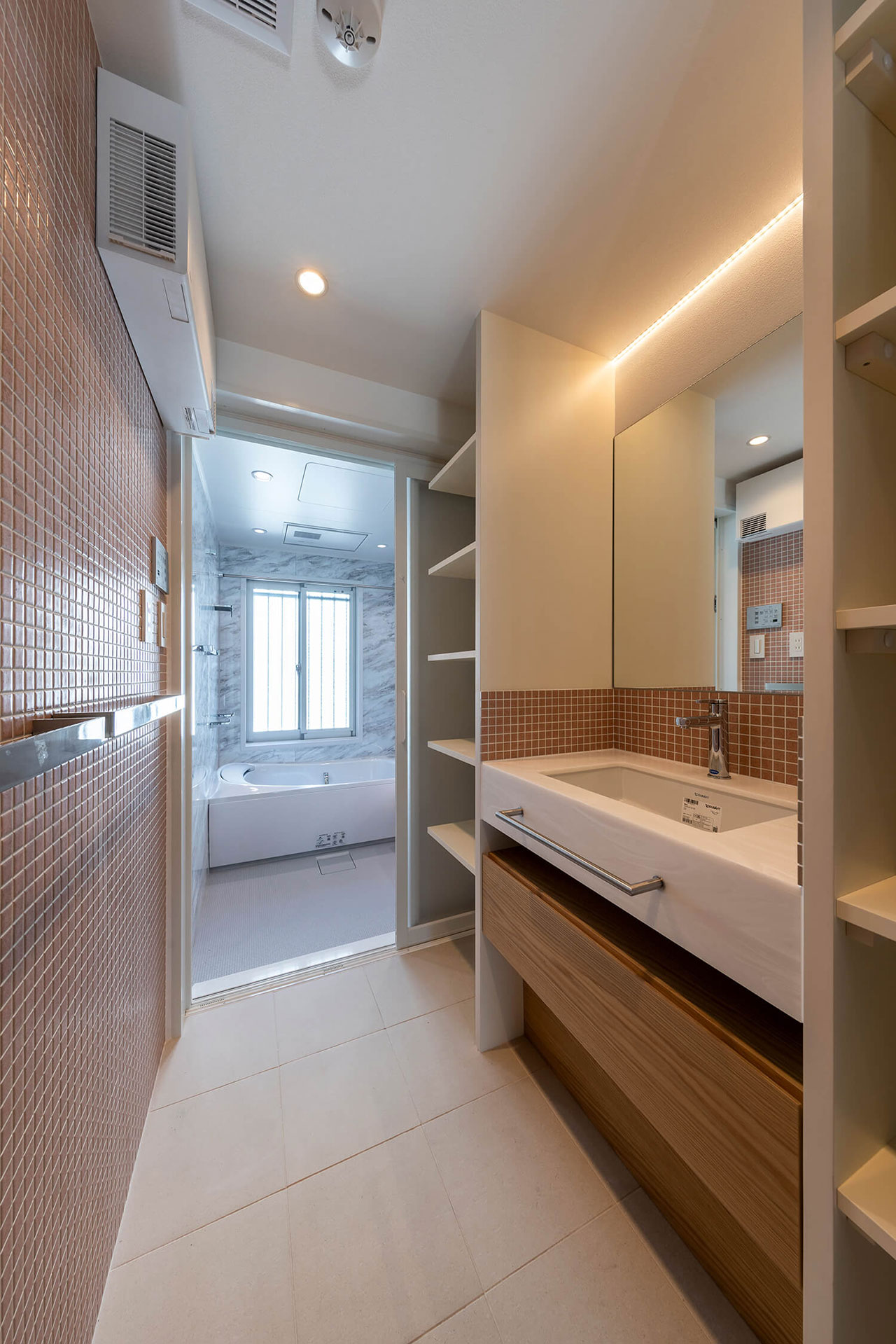 Modern bathroom with a sink, mirror, open shelving, and brown tile accents; a bathtub and window are visible in the adjacent room.