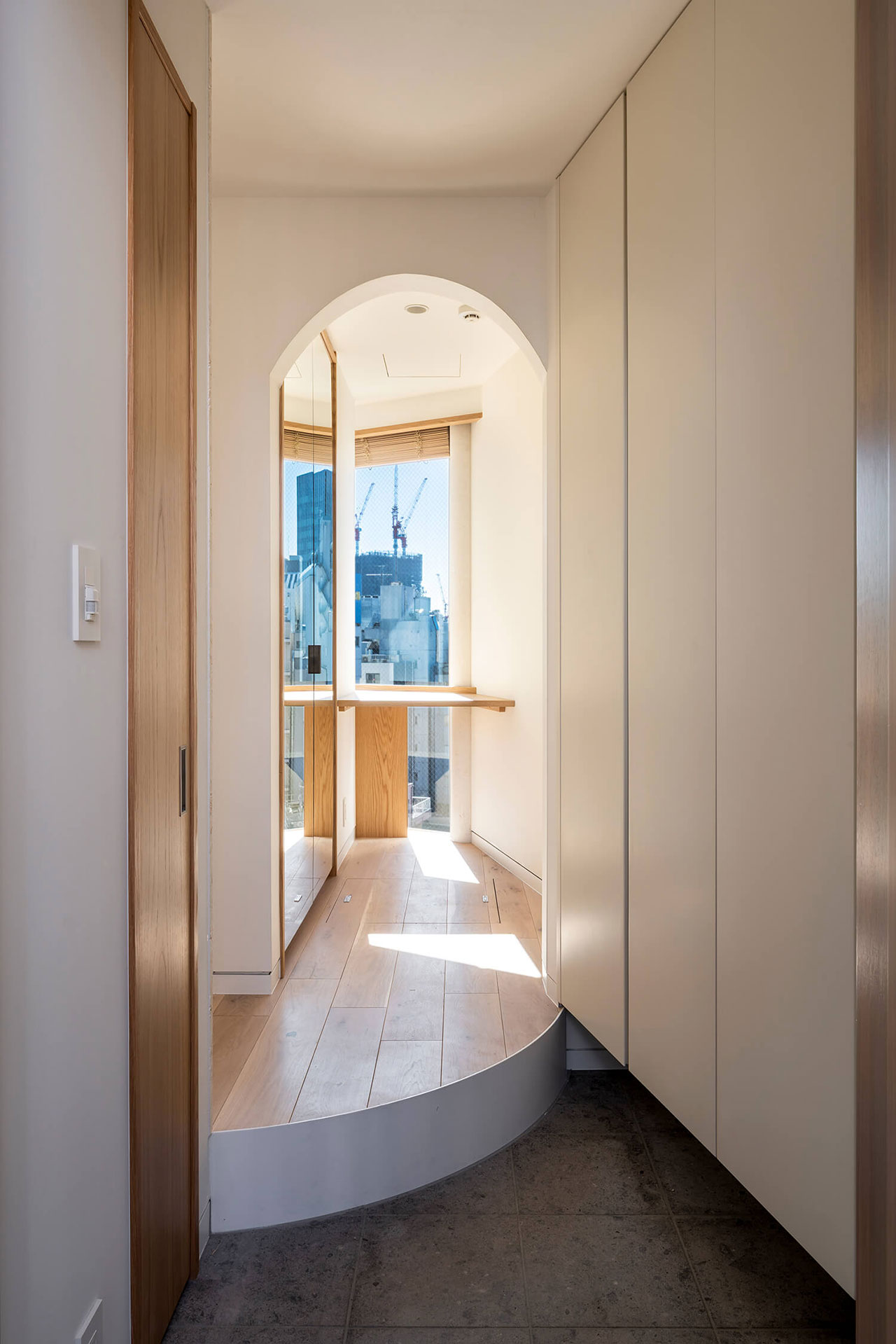A narrow hallway with white walls and built-in cabinets leads to a sunlit alcove with large windows overlooking a cityscape with cranes.
