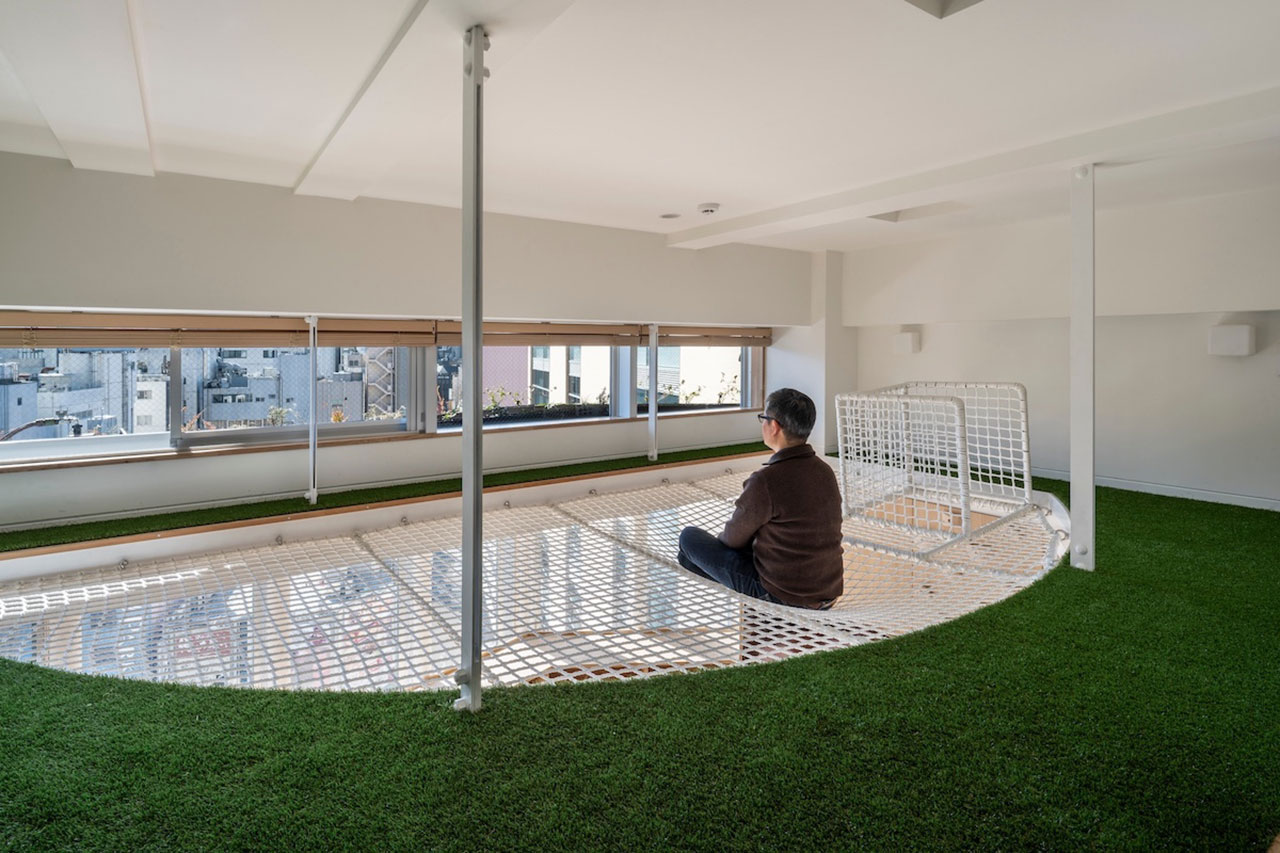 A person sits on a suspended white net platform in a bright room with artificial green grass flooring and large windows overlooking city buildings.