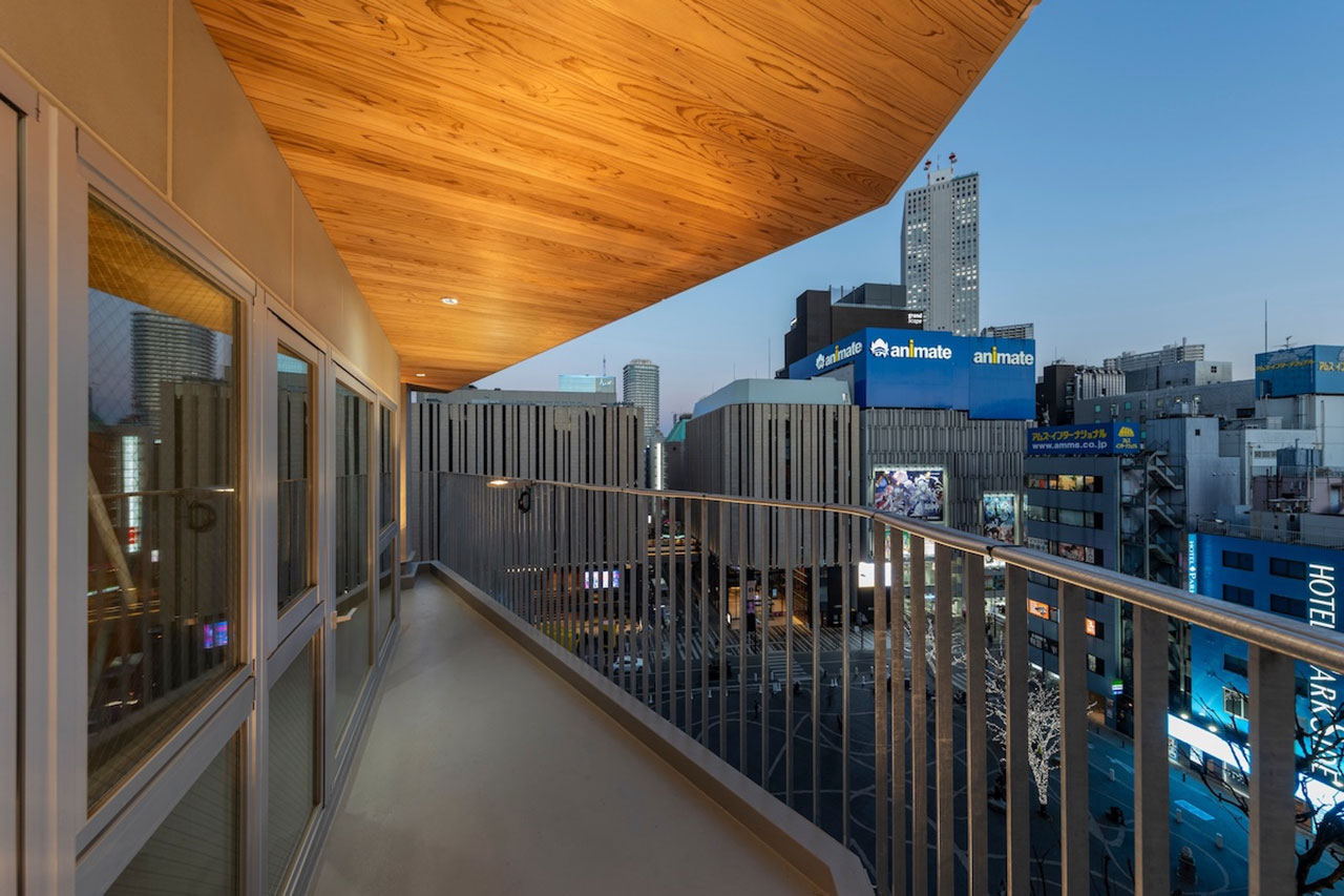 A modern balcony with a wooden ceiling overlooks a cityscape with tall buildings and illuminated signs at dusk.