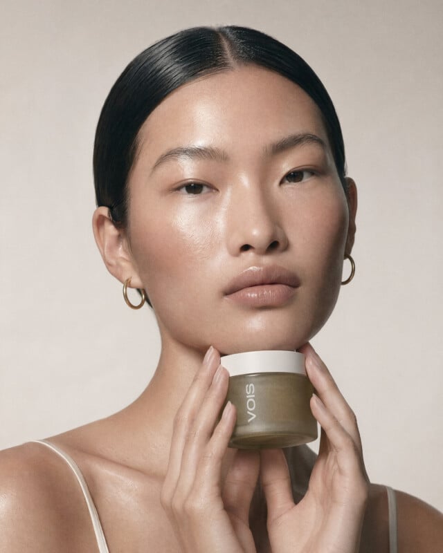 A woman with smooth, glowing skin and straight, dark hair pulled back holds a round jar labeled "Sioris" under her chin. She wears gold hoop earrings and a thin-strapped top, posing against a neutral background.