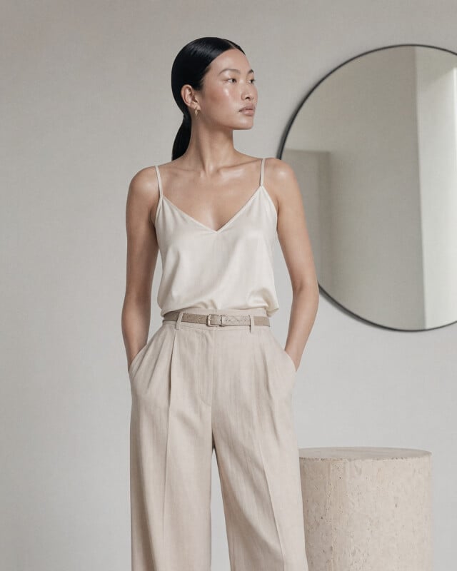 A woman in a cream satin camisole and high-waisted beige trousers stands with hands in her pockets. She has sleek black hair tied back and is looking to the side, with a round mirror on the wall behind her.