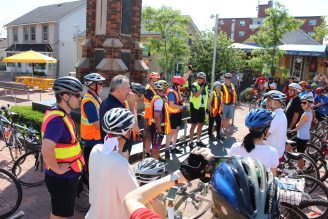 A group of cyclists wearing helmets and high-visibility vests gather with their bikes in a sunny public square for a community ride, listening to a speaker near a brick monument and nearby storefronts.
