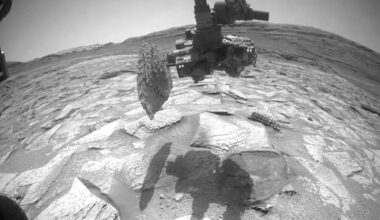 A robotic arm from a Mars rover holds a rock sample above a cracked, rocky Martian surface, with the rover's shadow visible on the ground and hills in the distant background.