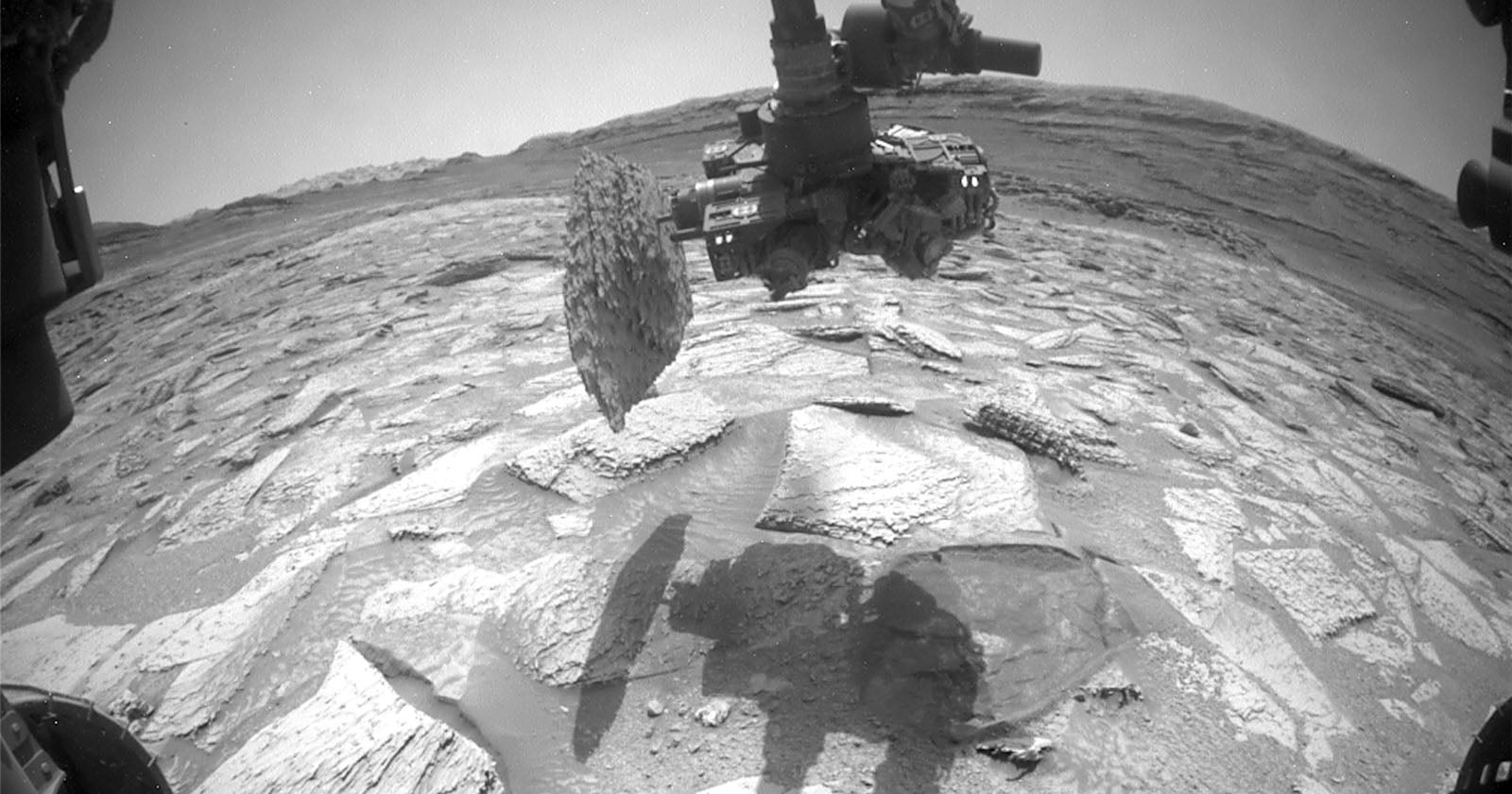A robotic arm from a Mars rover holds a rock sample above a cracked, rocky Martian surface, with the rover's shadow visible on the ground and hills in the distant background.