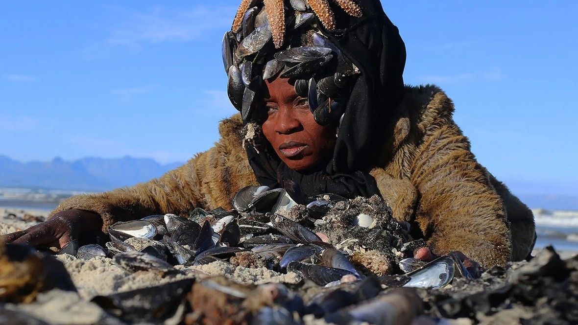 A woman wearing a fur coat and a black headscarf adorned with mussels and a starfish lies on a sandy beach behind a pile of seashells.
