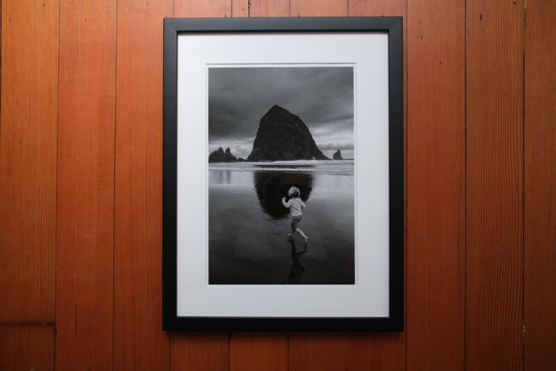 A framed black and white photo on a wooden wall shows a child running on a beach with Haystack Rock and its reflection in the wet sand under a dramatic, cloudy sky.