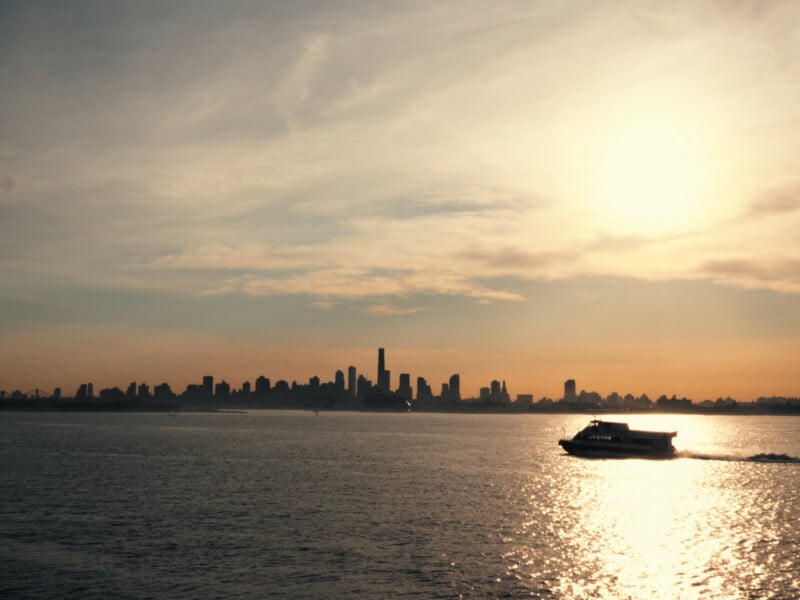 A boat glides across calm water at sunset, with the sun low in the sky and a city skyline silhouetted in the background against an orange and blue sky.