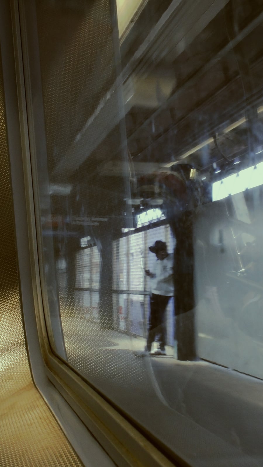 Blurred view through a train window showing a person standing on a station platform under overhead lights, with metallic and glass textures in the foreground.