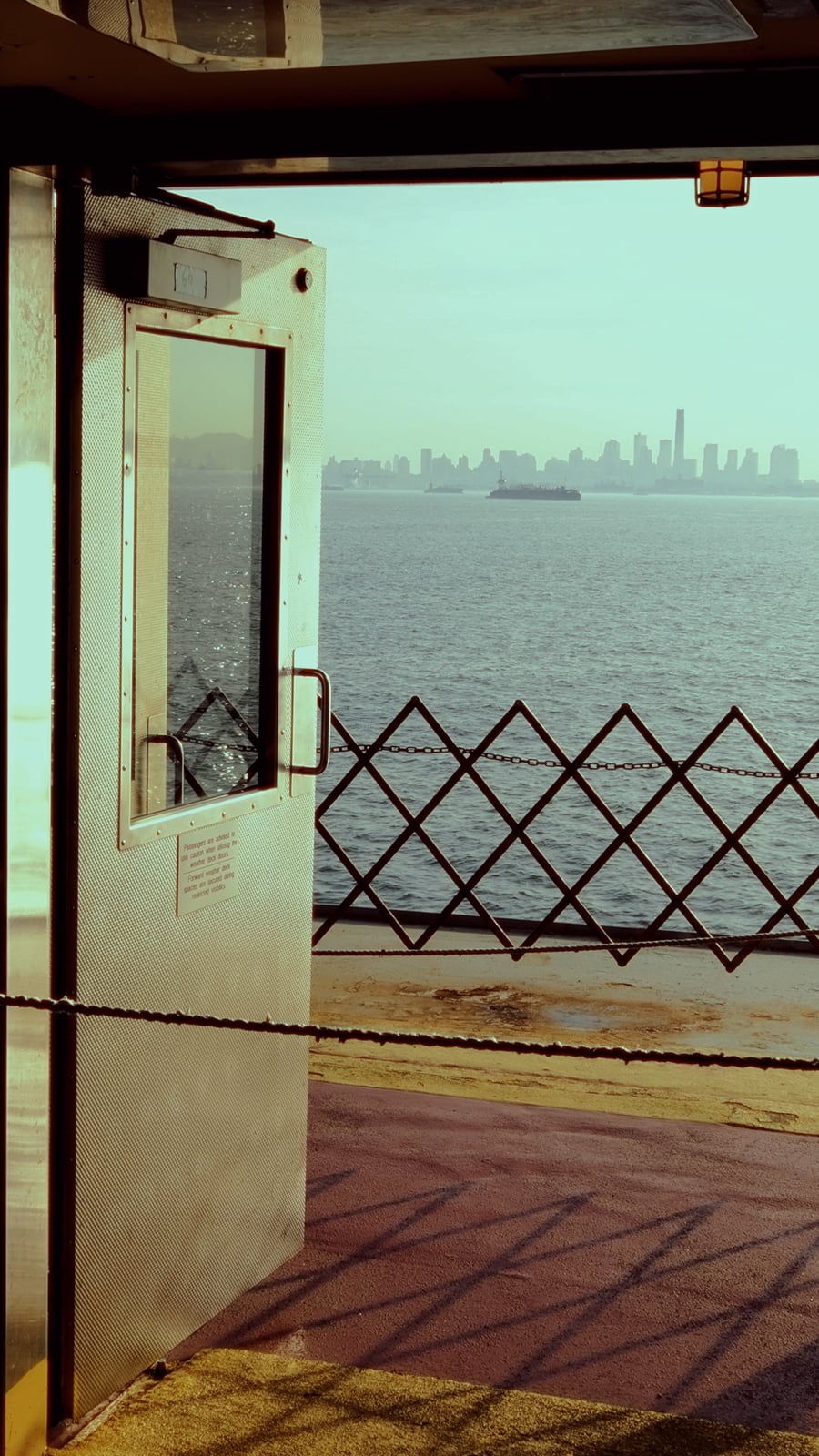 A metal door on a ferry opens to reveal a view of calm water and the distant city skyline under a hazy sky, with a safety barrier visible along the deck.