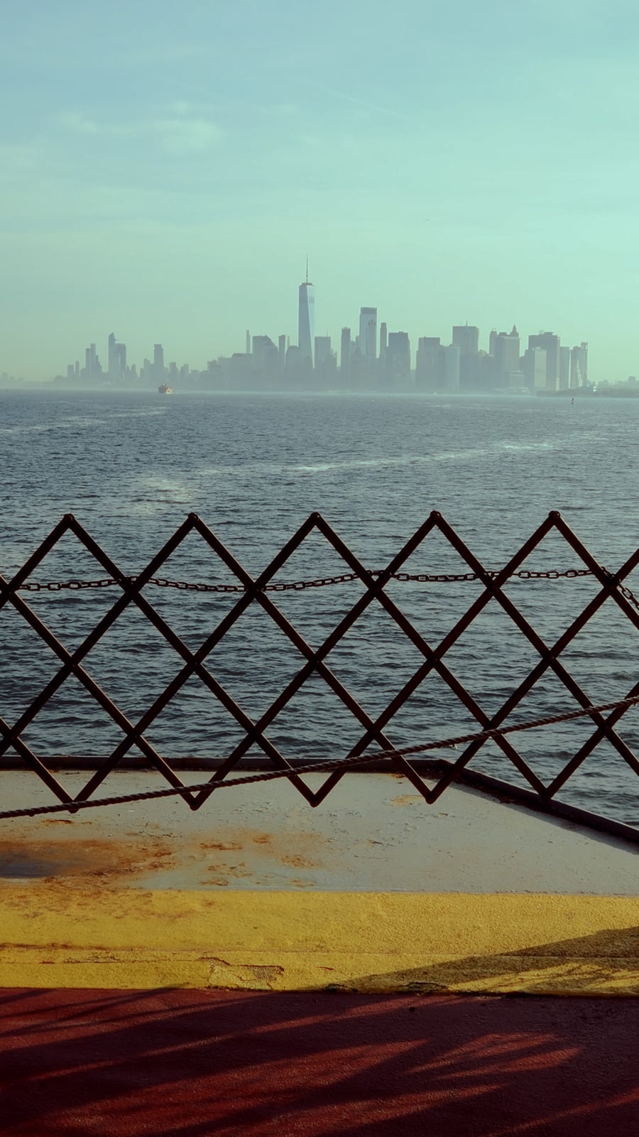 A hazy view of a city skyline across the water, seen from behind a ferry’s metal safety railing. The foreground includes yellow and red painted surfaces, while the city buildings rise in the background.