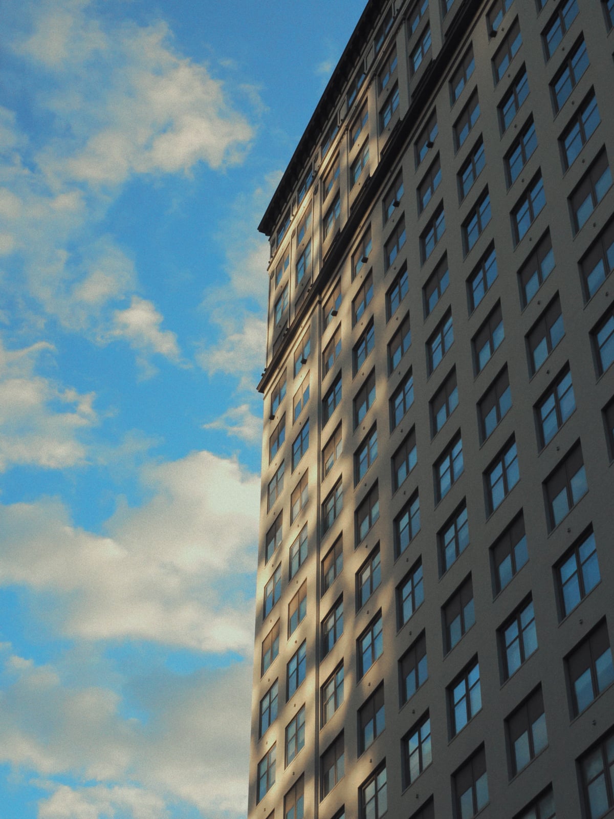 A tall building with many windows is shown from a low angle against a blue sky with scattered clouds, with sunlight casting shadows on the structure.