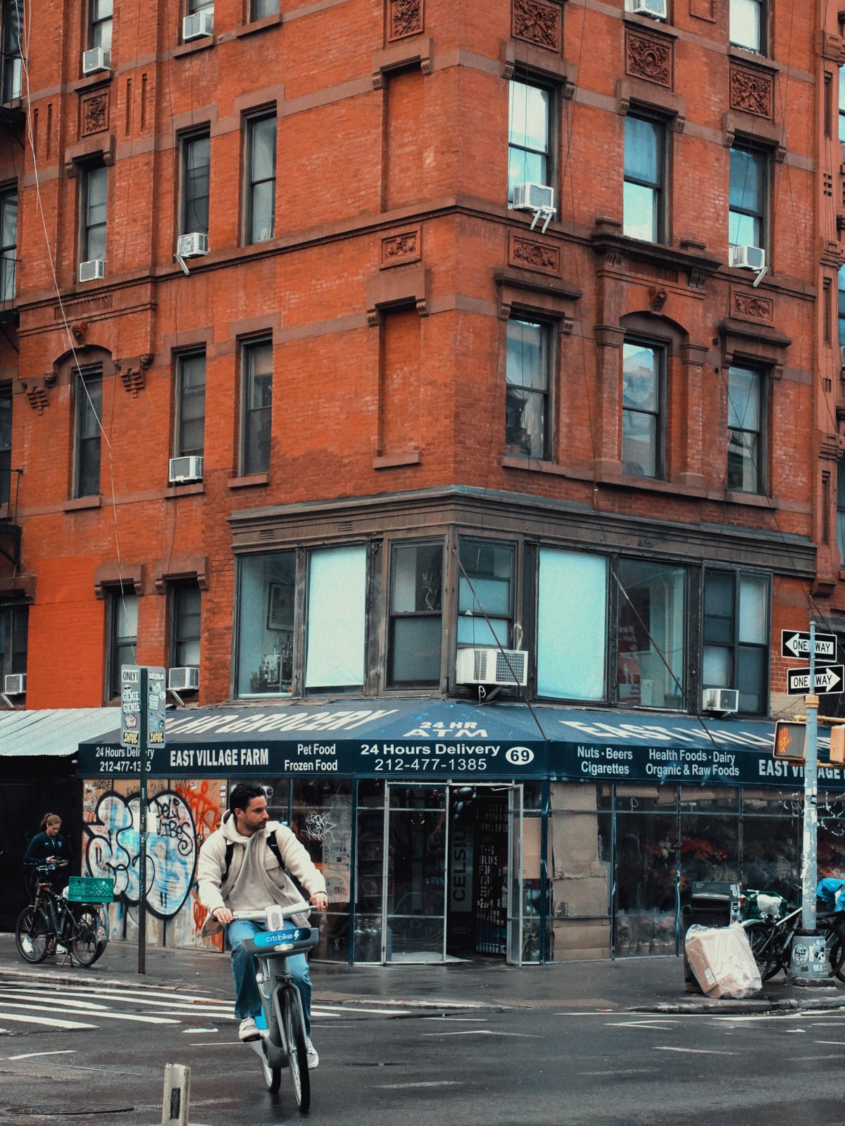 A man rides a bicycle across a wet street in front of a red brick building with shops at the corner, including “East Village Farm.” Other bikes are parked nearby, and the sky is overcast.