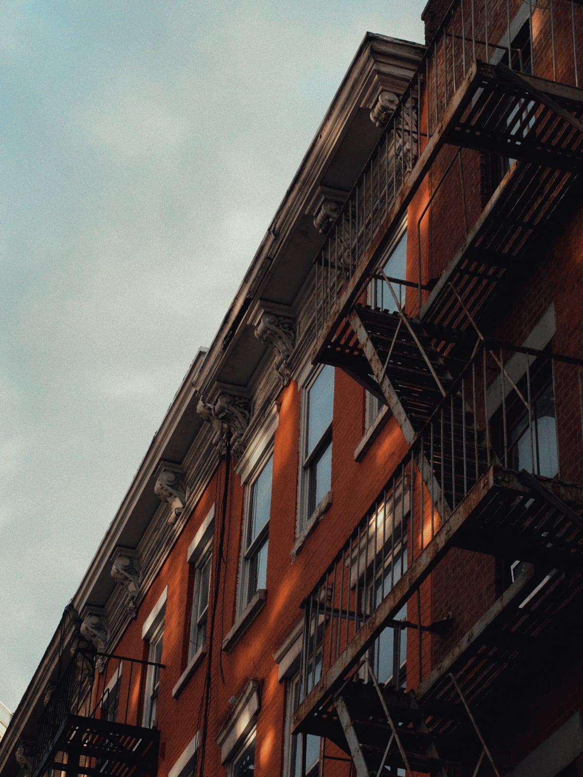 A brick apartment building with black metal fire escapes and white trim, photographed at an upward angle in warm afternoon light against a partly cloudy sky.