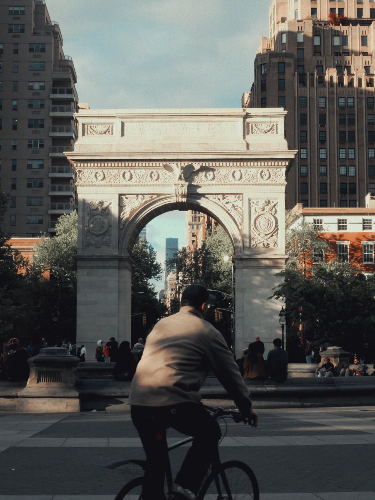 A person rides a bicycle in front of the Washington Square Arch in New York City, with tall buildings and trees in the background and people sitting and walking around the plaza.