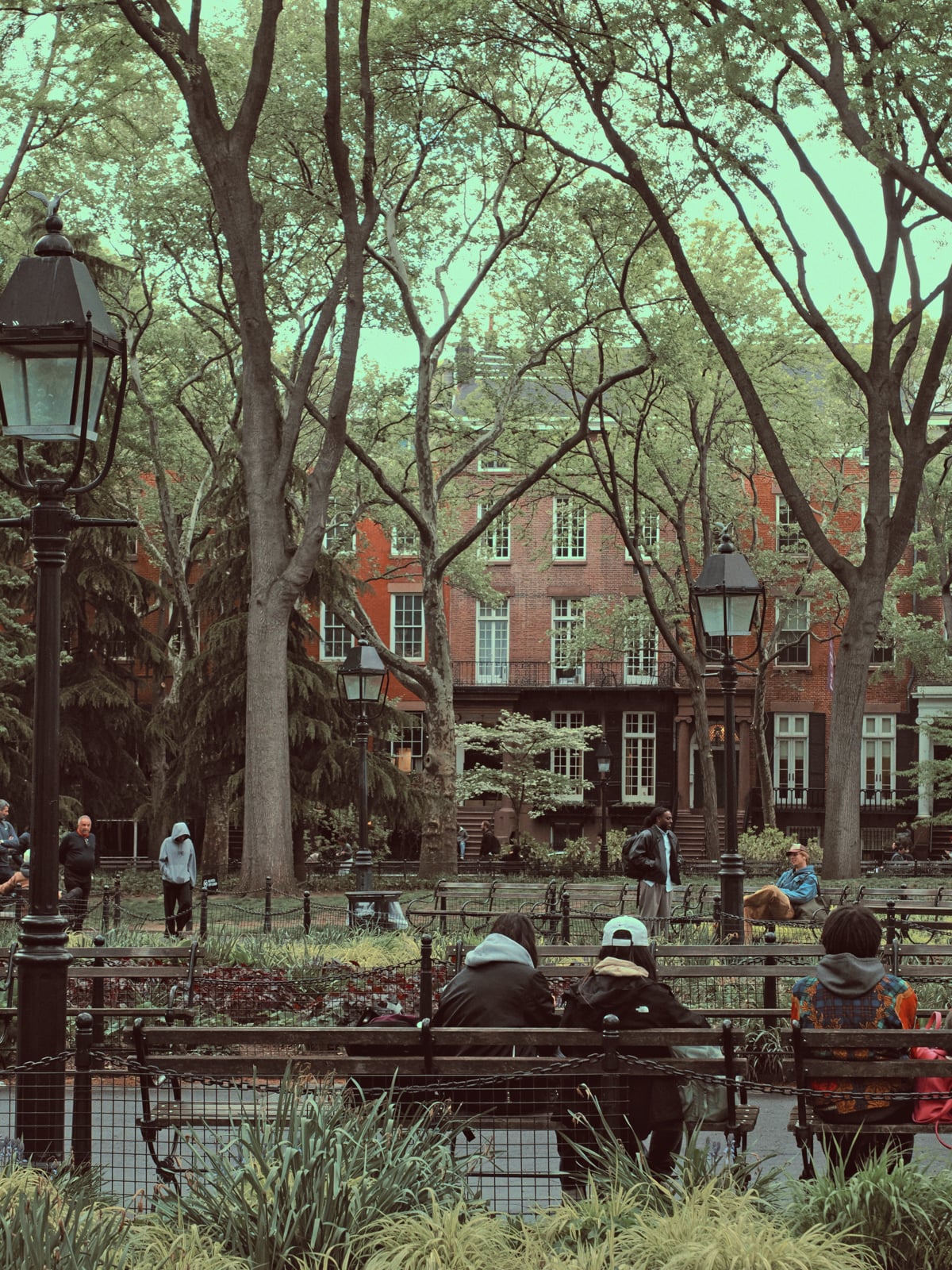 People sit on benches and walk through a park with tall trees and lampposts, surrounded by greenery. In the background, there are red brick buildings with large windows. The overall scene appears calm and peaceful.