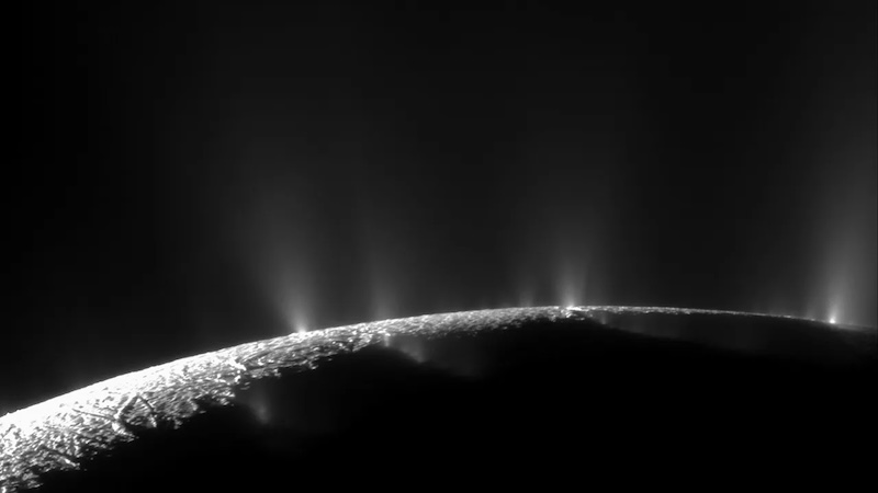 Sunlit edge of planet-like body, with geyser-like jets of vapor erupting upward against a black background.