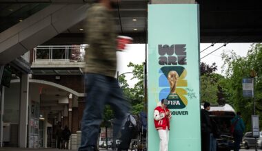 A person leans against a sign on a concrete post that reads, &lsquo;We Are FIFA Vancouver.&rsquo; A blurry pedestrian can be seen walking in the foreground.