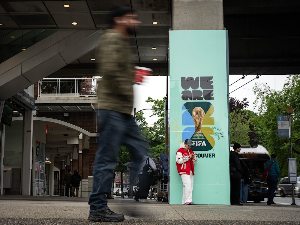 A person leans against a sign on a concrete post that reads, &lsquo;We Are FIFA Vancouver.&rsquo; A blurry pedestrian can be seen walking in the foreground.