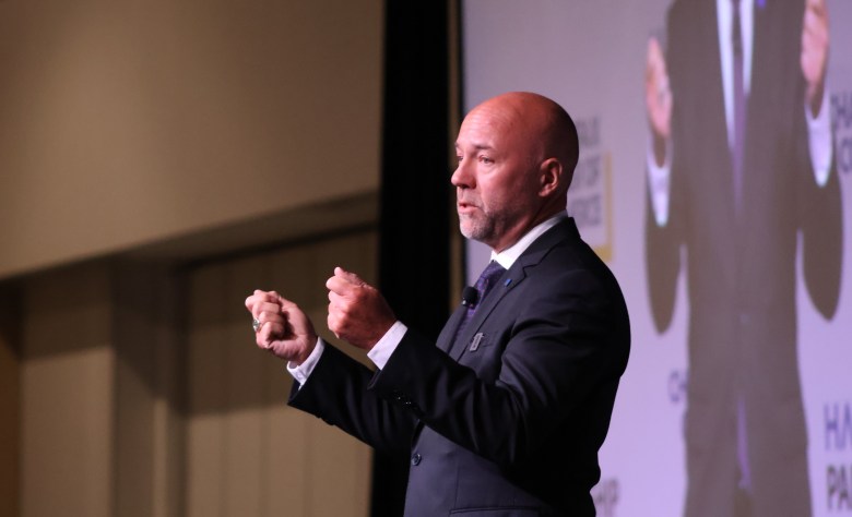 A white man with a shaved head and wearing a black suit with a white shirt, and patterned tie holds up his hands as he delivers a speech. Behind him is a video screen showing him delivering that speech.