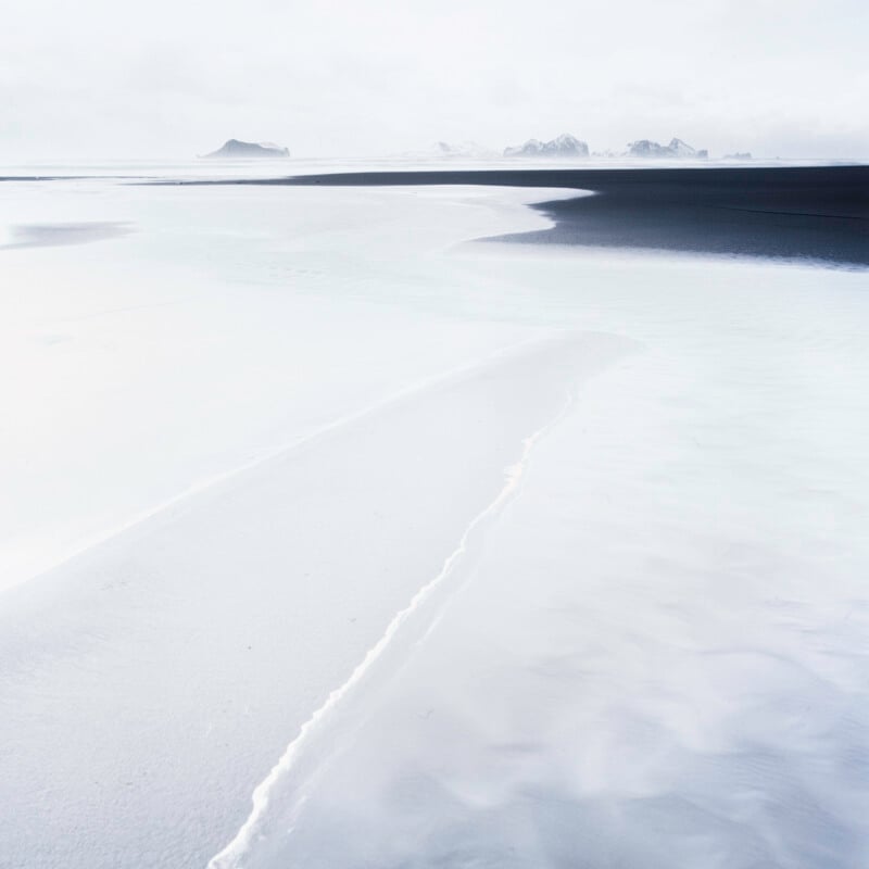 A minimal, icy landscape with pale, snow-covered ground, gentle curves, and distant mountains shrouded in mist under a bright, overcast sky.