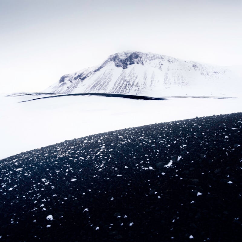 A snow-covered mountain rises in the distance under a cloudy sky, with a stark contrast between the black rocky foreground and the white, icy landscape beyond.