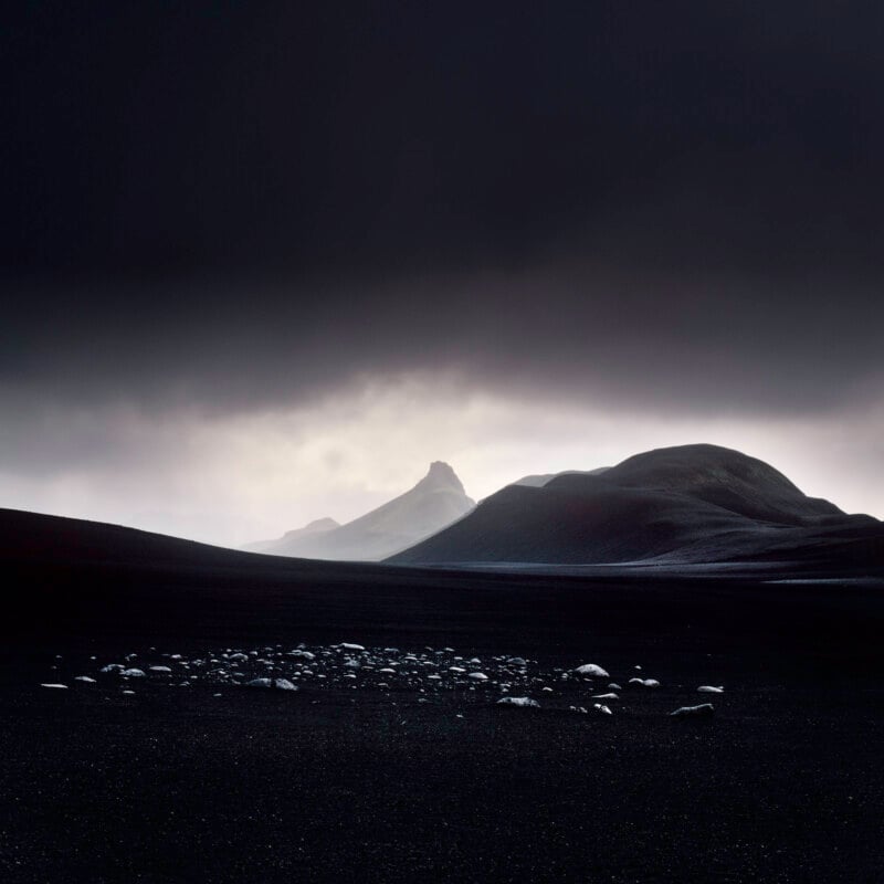 A dark, moody landscape with rocky hills under a cloudy sky. Light breaks through the clouds, illuminating a distant peak and casting soft light on scattered stones in the foreground.