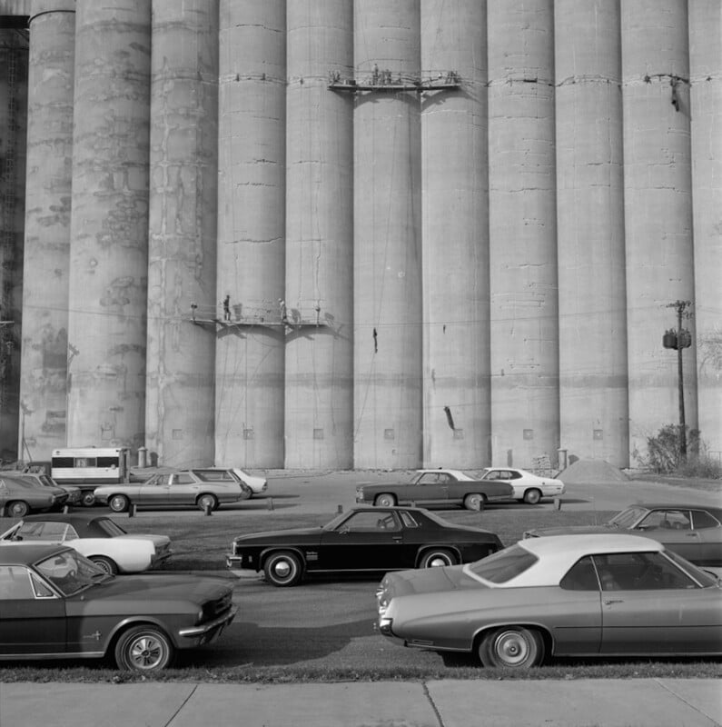 Several vintage cars are parked in front of tall, cylindrical concrete silos on a sunny day. The scene has a distinctly retro feel, likely from the 1970s or 1980s.