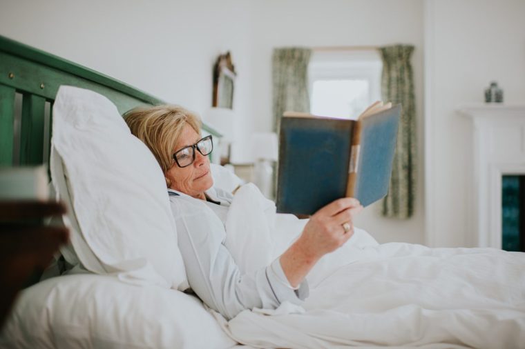 A mature woman lies in bed and reads a hardback book - stock photo