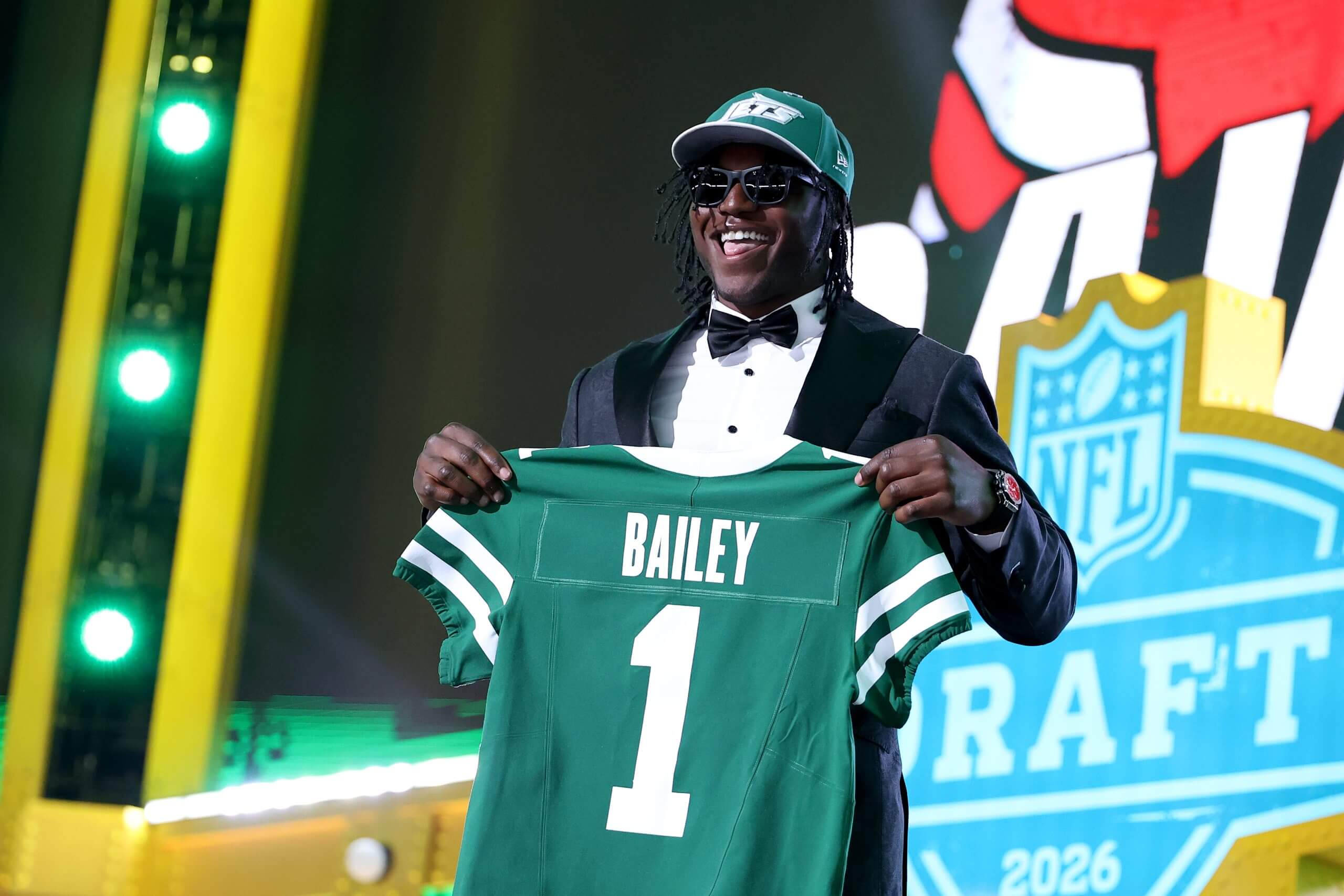 David Bailey, wearing a dark suit, black sunglasses and a green Jets hat, holds up a green No. 1 uniform while smiling wide.