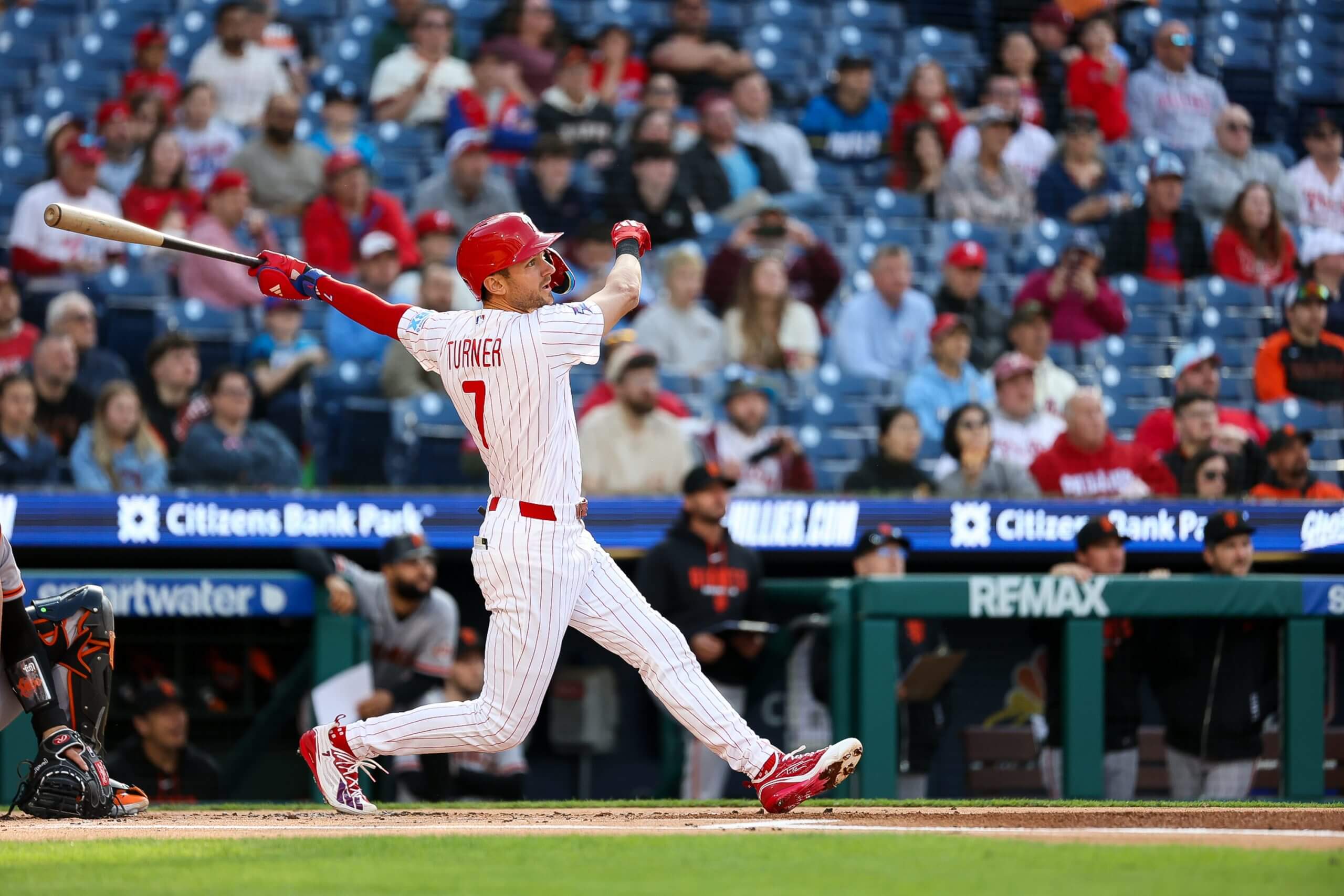 Trea Turner holds his bat up and behind him, watching the ball after a hit with the crowd in the background.