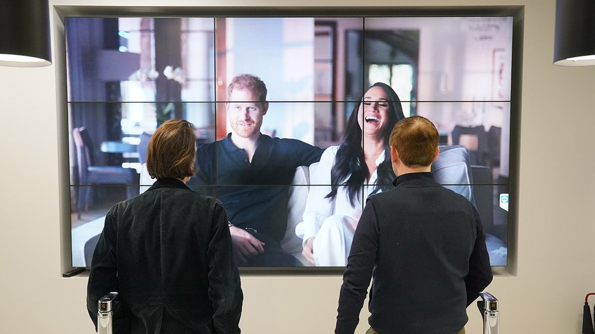 Two men watching the Duke and Duchess of Sussex documentary on a large screen in an office.