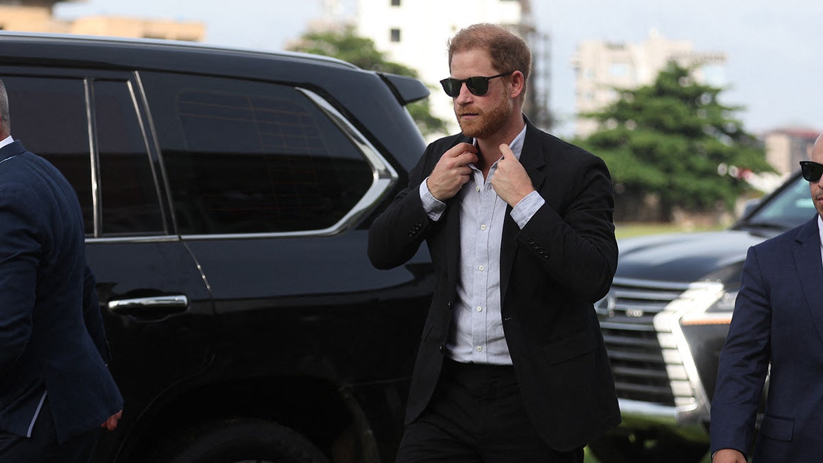 Prince Harry wearing sunglasses and fixing his tie next to a car at a polo club in Lagos