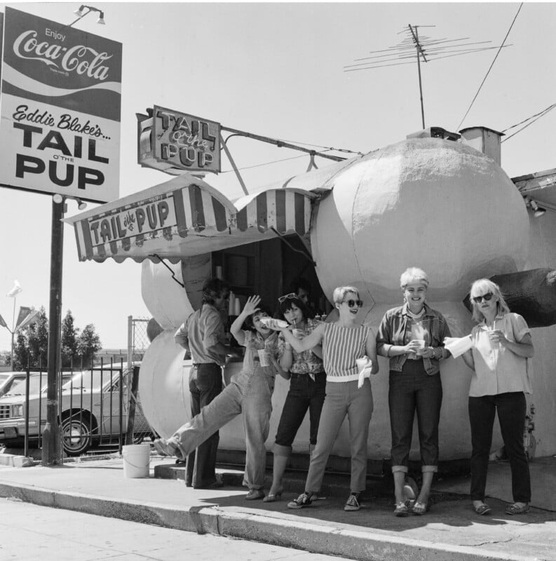 Five women pose and smile in front of the hot dog-shaped Tail o’ the Pup stand, with a large Coca-Cola sign above. Some wave, others hold food, and a car is parked beside the quirky building.