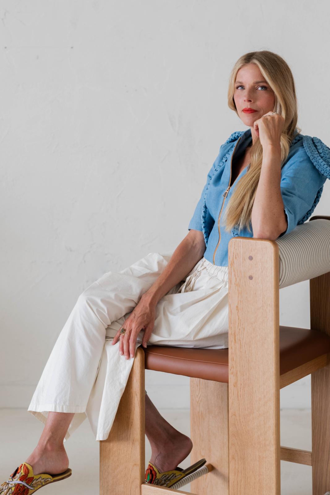 Portrait of a young blonde woman with long hair, dressed in casual-chic clothes in a studio while sitting on a wood and leather chair, or in a wood-clad office surrounded by books.