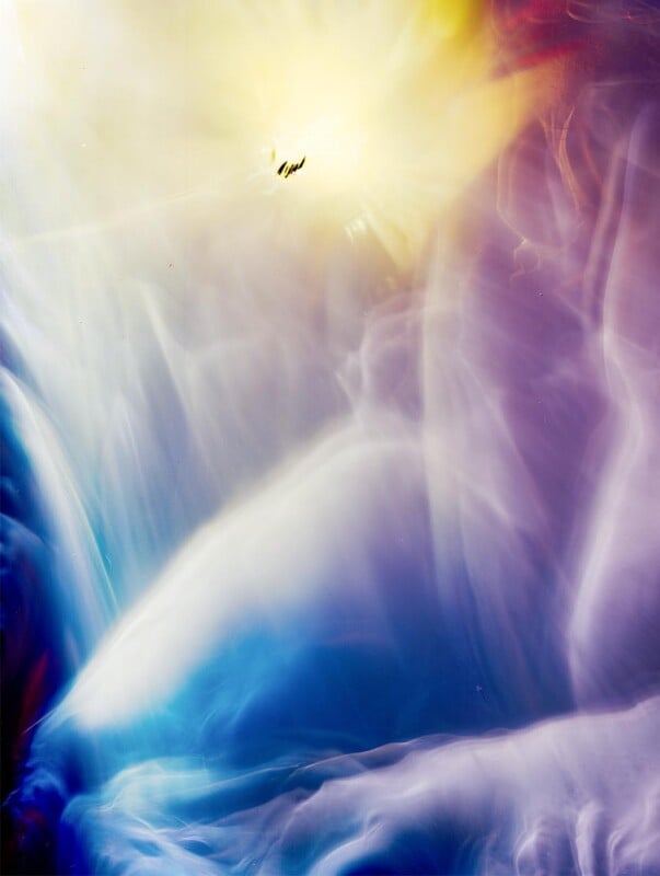 Abstract image with swirling, ethereal clouds in shades of blue, purple, and white, blending upwards into a bright yellow light. There is a small, dark silhouette near the center of the glowing area.
