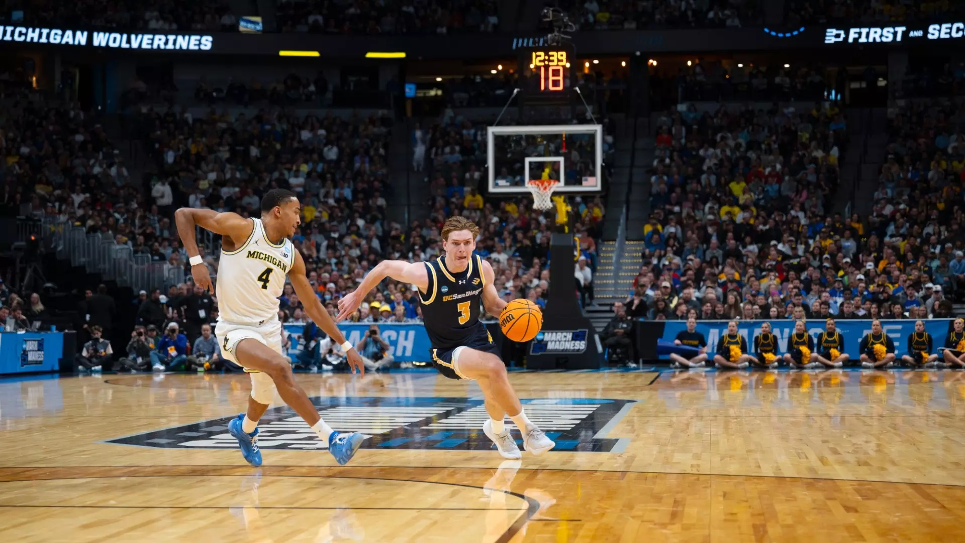 Wearing a blue jersey, Hayden Gray participates in the 2025 NCAA Men's Basketball Tournament, dribbling a ball against a Michigan defender