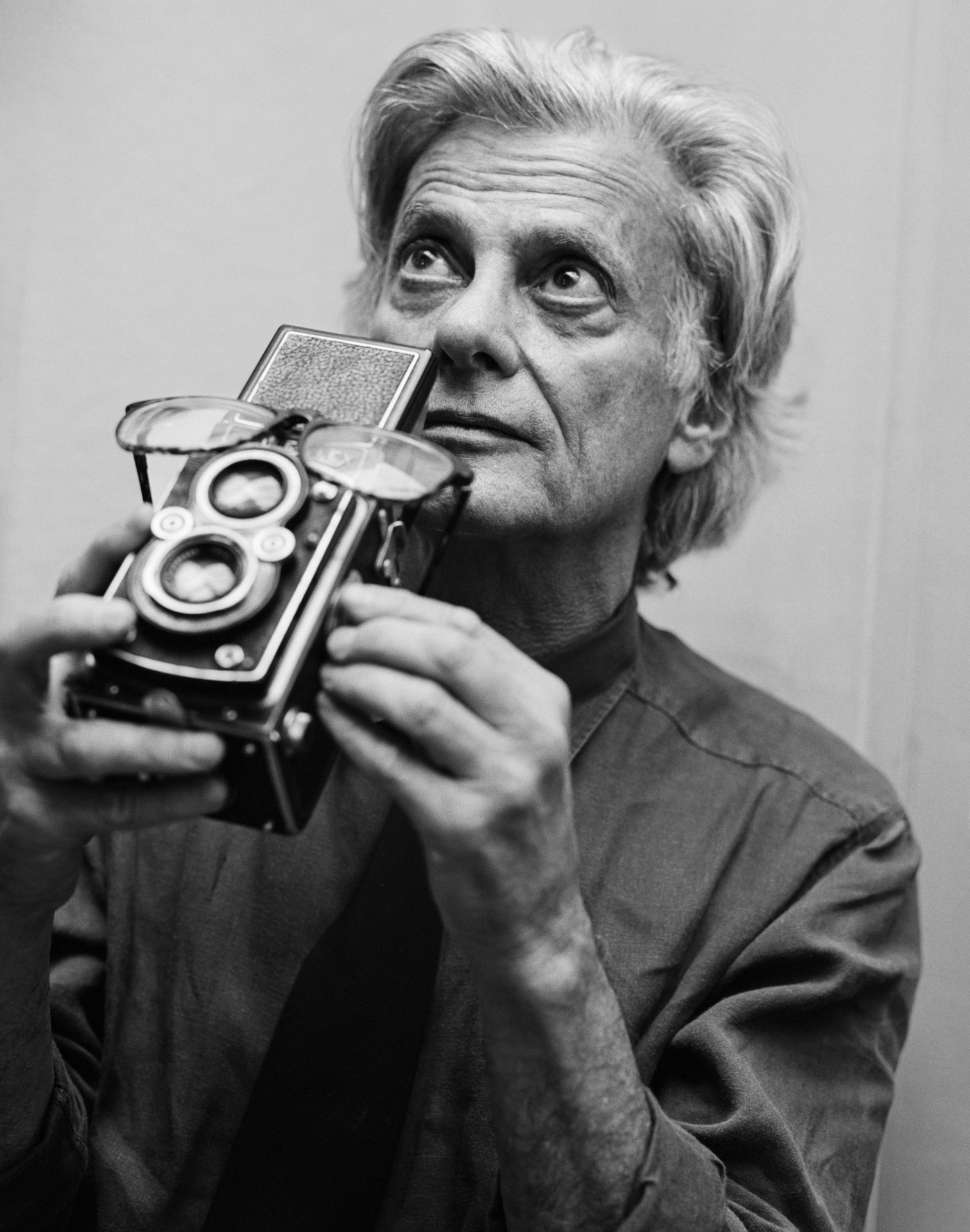 A black-and-white portrait of an older Avedon holding a twin-lens reflex camera up to his chin, his wide eyes fixed on something beyond the frame.