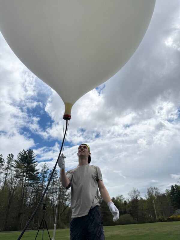 A person wearing a cap and gloves holds a large white balloon outdoors on a grassy field, with trees in the background and a partly cloudy sky above.