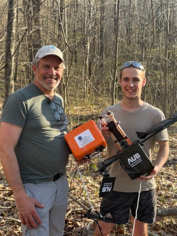 Two people stand in a forest, smiling at the camera. They are holding equipment, including an orange box and a black AUB-labeled device. Dry leaves cover the ground and bare trees fill the background.