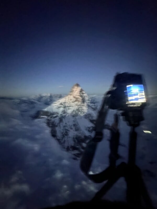 A blurred photo shows a camera on a tripod capturing a distant, snow-covered mountain peak at dusk or dawn, with a blue and purple sky in the background.