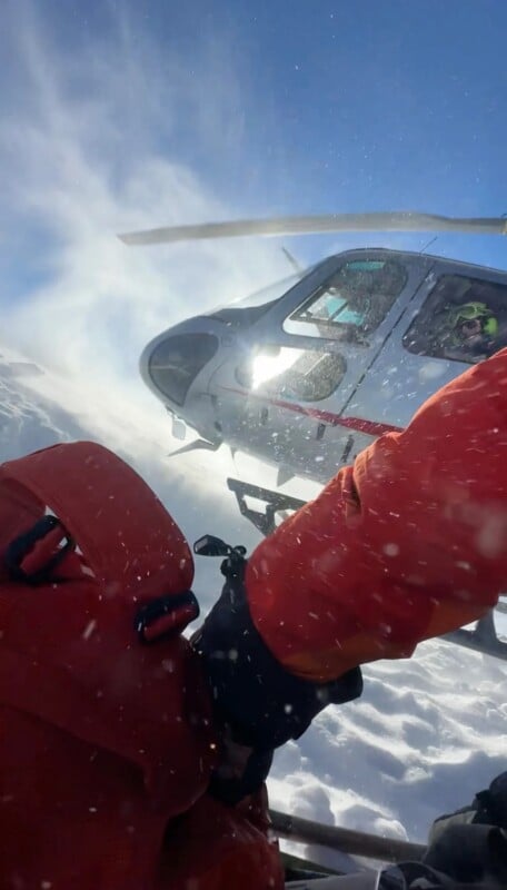 A helicopter hovers above snowy ground while a person in bright red winter gear moves through blowing snow, creating a dramatic scene in a mountainous, icy environment.