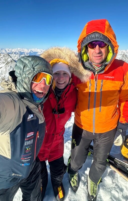 Three people dressed in heavy winter jackets and sunglasses smile on a snowy mountain peak with clear blue sky and distant mountain ranges in the background.