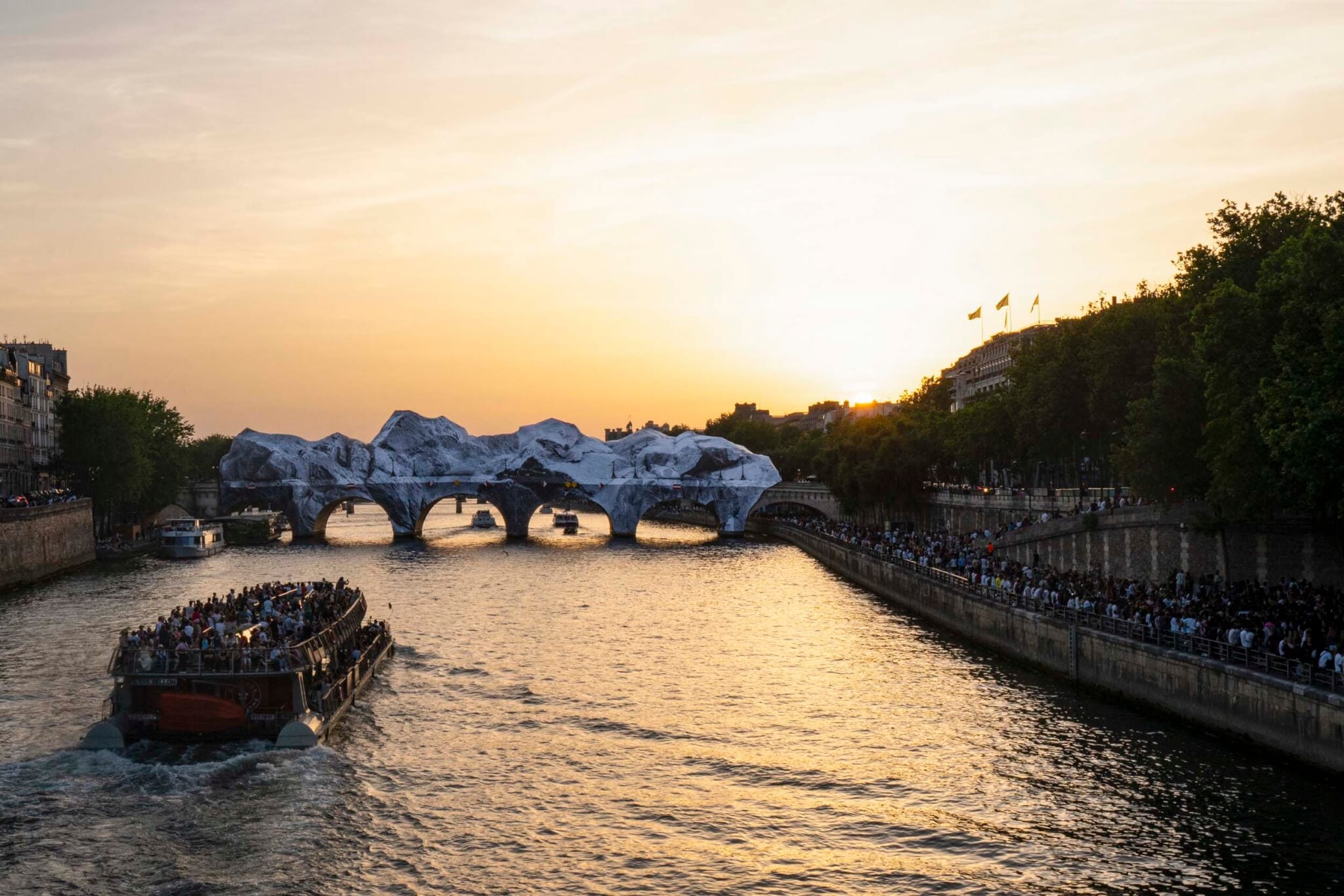 Large crowds line the embankment of the Seine at sunset, watching JR's mountain-shaped fabric installation cover the arches of the Pont Neuf