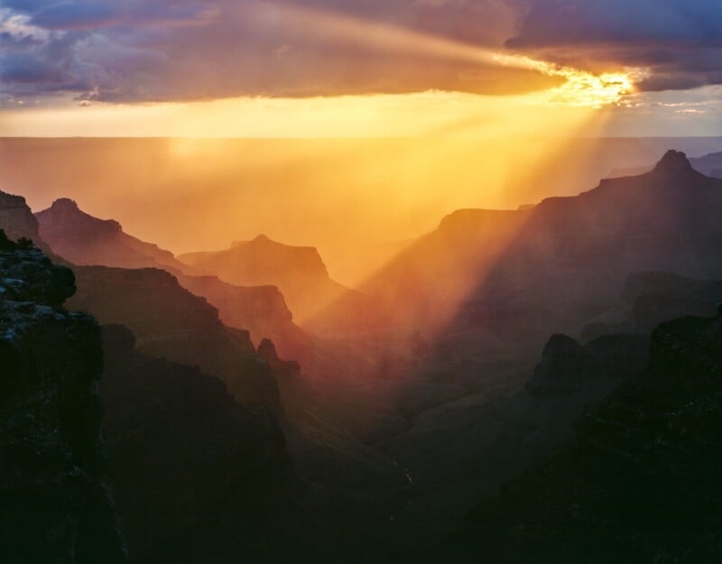Sun rays shine through clouds, illuminating the misty, layered rock formations of the Grand Canyon at sunset, creating a dramatic and colorful landscape scene.
