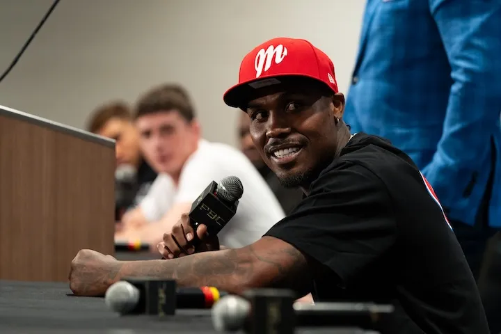 Jermall Charlo sits at a table at a press conference, holding a microphone in his right hand while his left forearm is rested on the table. Charlo is looking to the left in the direction of the camera, which shows him in side-profile.