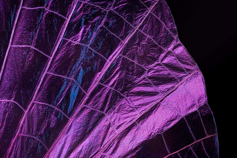 Close-up of a shiny, metallic purple surface with a textured, grid-like pattern and iridescent reflections, set against a dark background.